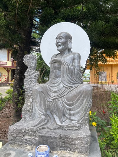 This outdoor image captures a large, grey stone statue within a temple complex in Đăk Mar, Vietnam. The central subject is a statue depicting an elderly, bald male figure with a wrinkled face, prominent ears, and a smiling expression. The figure is seated cross-legged, draped in flowing robes, with its hands in a gesturing posture. A large, plain white circular halo is positioned directly behind the figure's head. The statue rests on a dark, rough-hewn stone base. To the immediate left of the main statue, a smaller, vertically oriented grey stone carving features intricate, swirling patterns, possibly representing a stylized dragon or elemental motifs.
In the foreground, directly in front of the statue's base, a blue and white patterned porcelain incense burner containing dark ash is placed on a paved surface, flanked by two small white ceramic objects.
The background on the left features the trunk and dense green needles of a large evergreen tree. Beyond the tree, a multi-story building with traditional Vietnamese architecture is visible, characterized by yellow, red, and white elements, ornate curved rooflines, and a red decorative lantern.
The background on the right displays a large, golden-yellow temple building with tiered roofs and detailed architectural features. Scattered green foliage and shrubbery, including some with small yellow and red flowers, are present in the mid-ground. A smaller, lighter grey statue is partially visible in the distance to the right of the main subject. The scene is illuminated by clear daylight.