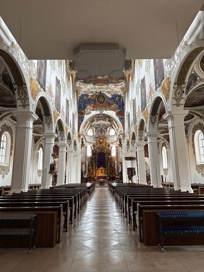 This image shows the interior of the baroque basilica of Saint Martin in Biberach an der Riß, Germany. The camera angle is at floor level, capturing the length of the nave. Rows of dark wooden pews line the right side of the image, leading toward the altar area. The aisle floor is tiled in a geometric pattern, reflecting light from the windows. On the left, ornate white columns and arches extend the length of the nave, supporting a highly decorated ceiling with religious paintings and frescoes. The architecture is characterized by elaborate details, typical of baroque style. The altar area is visible in the distance, featuring gold detailing and religious figures. No people are visible in the scene.