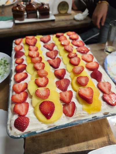 The image depicts a rectangular dessert in a glass baking dish, topped with rows of halved strawberries and circular slices of a yellow fruit, likely mango or peaches, arranged on a creamy white base. The dessert sits on a wooden surface, possibly a table or countertop. In the background, there are small brown ramekins arranged on a white serving platter, and a partial view of a person's arm and torso is visible on the upper right. A plate with chopped green ingredients is visible on the left side of the dessert. A glass of clear liquid and another plate are partially visible in the background on the right side. The overall scene suggests a home or restaurant setting where a dessert is being served or prepared. There is no indication that this image was taken in the city of Mosomishte, Bulgaria.