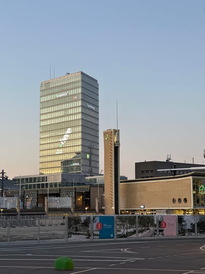 The image captures a modern urban landscape in Eindhoven, Netherlands, under the soft, muted light of either dusk or dawn. The scene is devoid of obvious human activity, contributing to a quiet atmosphere.
In the foreground, a wide paved area, possibly a road or plaza, features white painted lines. A small, artificial-looking green mound sits towards the right. Metal barriers or construction fencing line the edge of this area, displaying several large banners. One red banner clearly shows "Gemeente Eindhoven" (Municipality of Eindhoven) with a crest-like logo. Another blue banner reads "Eindhoven Engine" and "Kennis en kunde verbinden" (Connecting knowledge and expertise).
Beyond the foreground, a collection of buildings rises. To the right, a distinctive light brown building features three circular windows and a lit "2" sign, potentially indicating a station entrance. Adjacent to it, a tall, slender structure stands, capped with a large, illuminated clock face displaying Roman numerals and green hands. Further to the left and dominating the midground is a tall, modern skyscraper with a reflective glass facade. This building is adorned with various illuminated corporate logos and names, including "JLL," "broadwalk" (partially visible), "evee" (inverted), "fokus metals," and "tikkfood."
The sky above is clear, a soft blue that suggests the transition between day and night, with artificial lights already illuminating signs and clocks on the buildings. The overall impression is one of a contemporary city preparing for or concluding its daily activities.