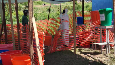 This outdoor image, set in Tchere, Democratic Republic of the Congo, depicts a makeshift hygiene or decontamination station under a tarp roof, supported by wooden posts. Orange plastic safety netting encloses parts of the area, creating distinct zones.

Two individuals are present. On the left, a person wearing a green uniform, a surgical mask, and a face shield stands near the netting. On the right, another individual is fully clad in a white hazmat suit with orange stripes and gloves, appearing to be engaged in an activity or adjusting their gear.

The ground is grassy, and the bright sunlight suggests it is daytime, casting clear shadows. In the background, there are trees and distant structures. Several colorful plastic buckets and containers are arranged throughout the station. On the left, multiple pink, blue, and orange buckets are on the ground. On the right, elevated on wooden stands, are three larger containers: a blue bucket, a red drum, and a green drum.

Visible text provides functional details: the blue bucket is labeled "SAVON" (Soap). The red drum has "DESHA BL..." written on its side, likely indicating a purpose related to decontamination. The green drum is clearly marked "CHLOREE 0,05 %" (0.05% chlorinated solution), with additional text including "par jour" (per day) and "24 heures" (24 hours), implying its use for disinfection. The combination of protective gear and various cleaning solutions indicates a public health intervention, possibly for disease prevention or control. A watermark "Joseph Kisuki" is present in the lower right corner.