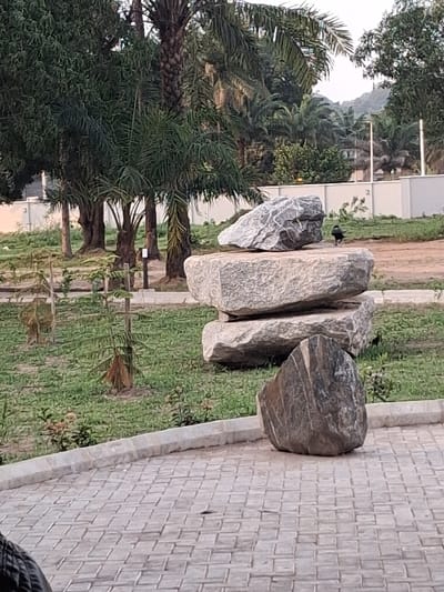 The scene depicts a landscaped outdoor area in Takum, Nigeria. In the immediate foreground, a grey interlocking paver surface is present, with a dark, indistinct object partially visible in the bottom-left. A large, dark, striated rock rests on this paved surface, right of center. Behind it, on a green grassy expanse, three lighter-toned, irregularly shaped rocks are stacked vertically. To the right of the stacked rocks, a single dark bird is positioned on the grass. The grassy area contains sparse vegetation and small planted saplings, bordered by a concrete curb separating it from the paved surface. The background is dominated by dense tropical foliage, including numerous palm trees and other broadleaf trees. A white perimeter wall is visible behind some of the foliage on the right, and faint, distant hills rise under a bright sky. No human subjects are present.