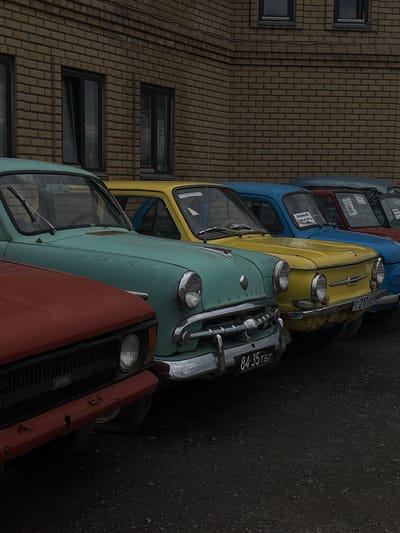A line of vintage cars is parked in front of a brick building in Bol'shoi Mashlyak, Russia. The cars are of various colors, including red, teal, yellow, blue, and dark gray. They appear to be old Soviet-era models. The building has several windows and a textured brick facade. The ground in front of the cars is dark and gravelly. The scene is overcast, with soft, diffused lighting. There are no people visible in the image.