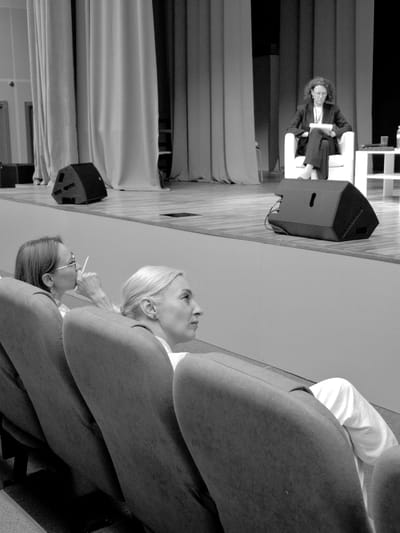 The black and white photograph, taken in Kungur, Russia, depicts a conference or presentation setting. Two women are the primary subjects. One woman, with curly hair and glasses, is seated in a chair on the left side of the frame, looking forward. She is wearing a dark jacket and pants. In the foreground, another woman with blonde hair is seen from the side, looking up and to the right. She is wearing a white top and a dark jacket. A third person's arm and shoulder, wearing a white sleeve, is visible in the upper right of the frame.
The scene includes a stage area with a podium or lectern on the left, and a large speaker is visible. Drapes or curtains hang in the background, creating a formal atmosphere. The lighting suggests an indoor event, possibly during the day. The expressions of the subjects appear attentive and engaged with the proceedings. There is no visible text in the image.