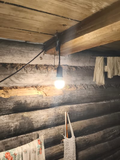 This image depicts the interior of a rustic wooden structure, likely a house or outbuilding, in Il'mino, Russia. The walls are constructed from rough-hewn logs, showing signs of age and weathering. The ceiling is made of unfinished wooden planks supported by beams.

A bare lightbulb hangs from a wire, illuminating the scene with a warm glow. To the right, several pieces of light-colored fabric are draped over a wire or beam, possibly drying clothes. In the lower left corner, a piece of fabric with a floral pattern is visible. On the right side of the image, a textured white bag with orange straps hangs from a nail. The logs themselves show gaps filled with what appears to be natural insulation, possibly moss or straw. The overall impression is one of a simple, functional living or storage space, typical of rural or older construction. There are no people visible in the image.