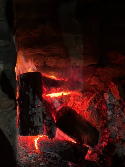 The image depicts a fireplace with burning logs. The logs are glowing red and orange, surrounded by ash and embers. The background appears to be the inside of a stone fireplace. No people, interactions, or other objects are visible. The environment is dimly lit. The location in Malak dol, Bulgaria is not relevant to the image content.