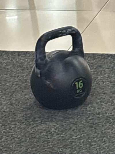 A black kettlebell with a weight marking of "16 KG" is positioned on a grey, textured carpet. The kettlebell is dark and appears to be made of cast iron. The handle of the kettlebell is curved and thick. Behind the kettlebell, a tiled floor is visible, with a subtle diagonal pattern of grout lines. The lighting suggests an indoor environment, possibly a gym or fitness area. The image is a close-up shot, focusing primarily on the kettlebell. There are no people visible in the image. There is no information in the image that would suggest the location is in Tri Protoka, Russia.