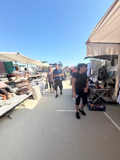The image depicts an outdoor market scene in Rakovski, Bulgaria, on a clear, sunny day. The market is lined with stalls displaying textiles, bedding, and possibly other household goods. Several people are visible walking through the market. In the foreground, a woman wearing a black polka-dot shirt and black capri pants is walking towards the right, holding a water bottle. Behind her, a man in a blue and white striped shirt and black shorts is walking, looking towards the camera. Further back, a couple is walking together, the woman in a floral print top and the man in a blue striped shirt. The stalls are covered by white awnings, and various items are neatly displayed. The ground is paved, and the overall atmosphere suggests a bustling marketplace.