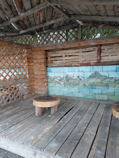 The image shows the interior of a rustic wooden gazebo located in the city of Ақши, Kazakhstan. The gazebo has a wooden plank floor and a wooden roof structure with exposed beams. The walls of the gazebo are partially constructed from logs, with one section featuring a lattice design made of wooden slats. Another wall is decorated with a mural depicting mountains and a body of water. A small, round wooden stool with three legs stands on the floor in the foreground. The environment appears to be natural, with greenery visible through the lattice work.