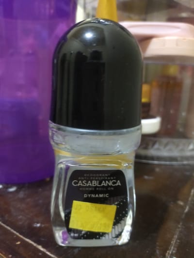 A close-up, eye-level, indoor shot shows a roll-on deodorant bottle with a black cap. The bottle has a clear glass base and a label with text that reads "DEODORANT ANTI PERSPIRANT CASABLANCA HOMME ROLL ON DYNAMIC". A yellow price sticker is affixed to the front of the bottle, obscuring some of the text. The bottle is placed on a wooden surface, possibly a table, with a blurry purple object to its left and other out-of-focus objects in the background. There are no people or animals visible in the image. The lighting is dim. The specific location in the city of Payacut, Indonesia, cannot be determined from the image alone.