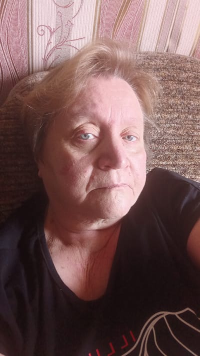 The image shows a close-up, selfie-style photograph of an individual who appears to be a mature woman with fair skin and light brown hair. She is wearing a dark t-shirt. The background consists of a textured, patterned surface, possibly wallpaper, in shades of beige and pink, and a textured brown surface that could be upholstery or fabric. The lighting suggests an indoor setting. The subject's expression is neutral, and she is looking directly at the camera. There is no visible indication of the city of Buy, Russia, within the image itself.