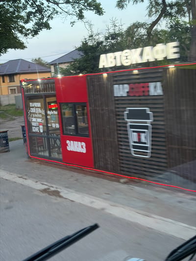 The image shows a roadside kiosk with the sign "АВТОКАФЕ" (Autocafe) in large white letters above. Below this sign, on a wooden slatted background, is a large graphic of a coffee cup with red and white accents. To the left of the coffee cup graphic, on the red side of the kiosk, the word "ЗАКАЗ" (Order) is written in large white letters. The kiosk has large glass windows, one of which displays text in Russian including "НАЧНИ СВОЙ ДЕНЬ С УЛЫБКИ" (Start your day with a smile) and "НАШЕГО КОФЕ" (Our coffee). The kiosk appears to be situated in an urban or suburban setting, with buildings and trees visible in the background. There are also two trash bins near the kiosk. The scene is captured from a slightly elevated, moving perspective, suggesting it was taken from a vehicle.