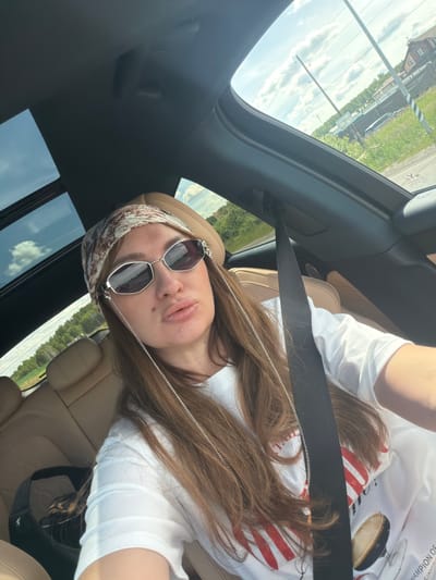 A woman with long brown hair is sitting in the passenger seat of a car, looking at the camera. She is wearing a white t-shirt, sunglasses, and a patterned headscarf. The car has a sunroof open, revealing a cloudy sky and a suburban landscape with houses, trees, and power lines. The interior of the car features tan leather seats and a black seatbelt. A black bag with a leopard-print lining is visible on the seat next to her. The scene appears to be taking place in Malinovka, Russia, given the context.