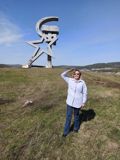 The image shows a landscape with a woman posing next to a large monument in Сатка, Russia. The monument consists of a Cyrillic letter "U" and a five-pointed star, painted in a metallic gray color. The woman is wearing sunglasses, a light blue jacket, and jeans. She is raising her hand in a waving gesture. The background includes a blue sky and a hillside with a town visible in the distance. The ground is covered in grass and dry vegetation. The image appears to be taken on a sunny day.