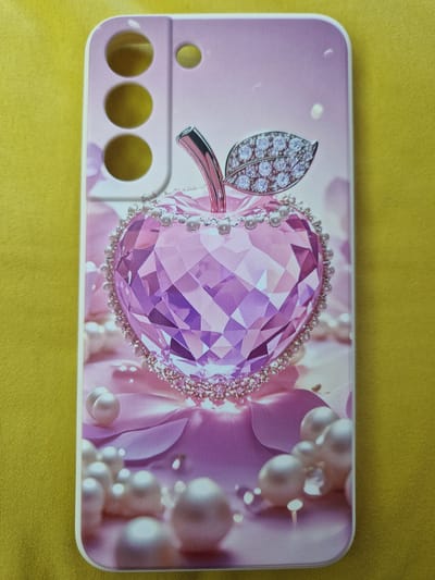 The image shows a close-up of a phone case with a pink and purple heart-shaped diamond apple design. The phone case is placed on a yellow textured surface, likely a fabric. The design features a large, faceted heart-shaped gem in shades of pink and purple, resembling a stylized apple. It is adorned with a pearl and rhinestone border. A silver stem and a diamond-encrusted leaf extend from the top of the gem. Scattered pearls and other sparkling elements are visible around the central design, against a soft, blurred background of pink and purple petals and light. The phone case has cutouts for a camera on the upper left side. The image does not provide any information or visual cues that would allow for the identification of its location in the city of Fresnes, France.