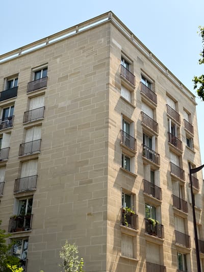This media file shows a view looking up at a building facade in Neuilly-sur-Seine, France. The building is constructed of light-colored stone blocks laid in a regular pattern. The facade is punctuated by numerous windows, many of which are fitted with dark metal shutters that are partially open. Some of these shutters have small potted plants attached, with green foliage visible. The building appears to be residential or office space, and the perspective suggests it is a tall structure. The sky is clear and pale blue, indicating it is daytime. The sunlight casts shadows on the facade, highlighting the texture of the stone and the depth of the window frames. The time of day appears to be morning or afternoon, judging by the angle of the light. The presence of trees with green leaves at the top and on the right side of the frame suggests a mild or warm season. There is no visible text in the image.