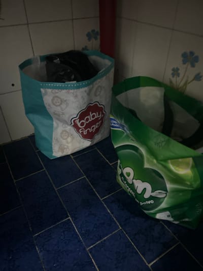 The image shows a corner in a room, likely a laundry area. There are two open bags on the floor. One bag is green and white, labeled with the brand "OM" detergent. The other bag is light blue and white, with a logo that says "Baby Finger". The floor is tiled with blue square tiles on one side and white square tiles on the other. The location is unspecified, and there is no information about people, actions, or interactions. The image is a still photograph of a domestic interior setting.