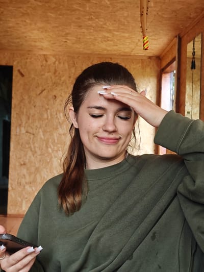 A young woman with brown hair pulled back in a ponytail, wearing a green sweatshirt, stands indoors. Her eyes are closed, and she is touching her forehead with her right hand. She is holding a black smartphone in her left hand. The background consists of wooden walls and ceiling. The image shows a portion of a building's interior, possibly a workshop or unfinished room, given the exposed wood paneling. There is a reflection of a window and possibly a door in the background on the right. A baseball bat with yellow and red tape is hanging from the ceiling. This scene is located in Kolyvan, Russia.