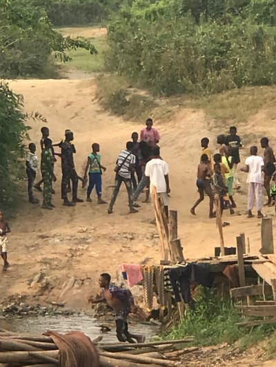 A group of people is walking on a dirt road between trees. The group consists of men and women dressed in various clothes. Some are wearing light-colored clothing, while others are wearing darker colors. Some have their backs turned, and some are facing the camera. There is a wooden structure nearby and some piles of wood.