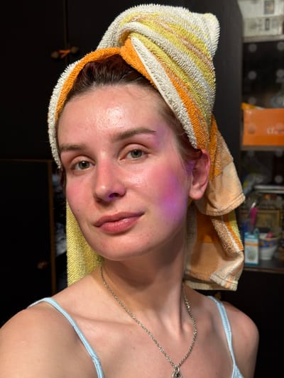 The image presents a close-up, rotated portrait of a young person, likely female, captured indoors in Заболотово, Russia. Their head is wrapped in a striped towel, predominantly white, yellow, and orange, suggesting recently washed hair or post-shower activity. The individual looks directly into the camera with a neutral expression.
A distinctive feature is a vibrant pinkish-purple glow visible on the left side of their face, encompassing the cheek and temple area, and subtly inside the left nostril. Their skin appears dewy or moist. They have green or hazel eyes and naturally colored, slightly parted lips. Around their neck, a delicate silver chain necklace is visible. The person is wearing a light blue strap from what appears to be a camisole or similar top. The background is dark, out of focus, and suggests an indoor setting with indistinct objects or shelving in the dimly lit environment. No visible text is present.