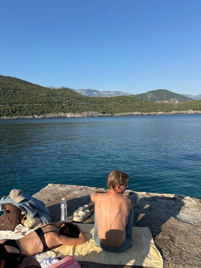 The image captures a serene scene at Dubraljevina, Montenegro, under a clear blue sky, suggesting a warm, sunny day, likely midday. Two individuals are relaxing on a concrete structure by the water.
In the foreground, a woman is lying on a yellow towel, wearing a black bikini top and resting her head on her hands. Beside her is a partially visible person, also seemingly relaxing. On the concrete, near their feet, are various items including a bottle of "VODAVODA" mineral water, a pair of beige Crocs, a checkered shirt, and a straw hat.
Further into the scene, a man with blonde hair, shirtless and wearing sunglasses, is lying on another towel, facing away from the camera, looking out at the vast expanse of the blue sea. His body is illuminated by the sun, casting a distinct shadow on the concrete.
The background features a dramatic landscape of steep, verdant hills that meet the coastline. The sea is calm with gentle ripples, reflecting the bright sunlight. The overall mood is one of peaceful relaxation and enjoyment of the natural beauty of the coastal environment.