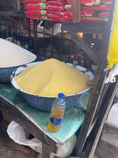 The image shows a cluttered market stall in Ojodu, Nigeria. On display are bags of food items, including cornmeal, rice, and packaged goods like Golden Penny pasta. Other items present include cooking oil in bottles, plastic containers, a broom, and other miscellaneous supplies. The stall is constructed of weathered wood and metal mesh, with various bags and containers serving as storage. The scene suggests a bustling, if somewhat disorganized, retail environment typical of an open-air market.