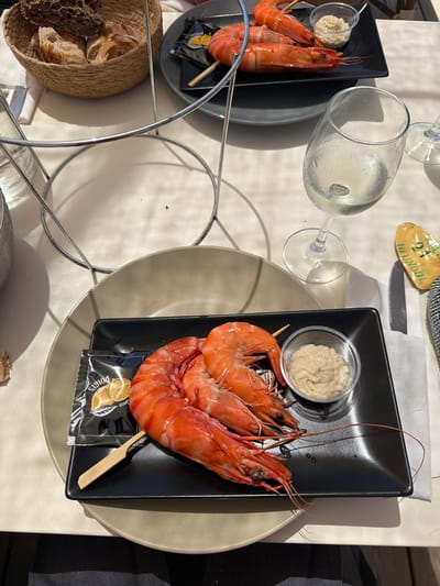 The image depicts an outdoor dining setting, likely on a sunny day, with a focus on a meal of prawns. In the foreground, a black square plate holds three large, cooked prawns on a skewer, accompanied by a small dish of creamy sauce and a packet of seasoning, possibly with a lemon graphic. A wooden skewer pierces the prawns. To the right of the plate, a nearly full glass of white wine with condensation sits on a table. Further to the right, a small, yellow object with green writing, possibly a decorative item or a souvenir, is visible. In the background, a woven basket contains slices of bread, and another plate with prawns and a dipping sauce is visible, suggesting multiple servings or dishes. The overall setting appears to be a restaurant in Lège-Cap-Ferret, France, given the context of the provided information. The table surface is white, and shadows are cast from sunlight, indicating an outdoor or well-lit environment.