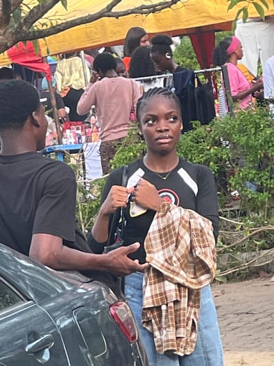 The image, taken in Ijanikin, Nigeria, captures a street scene with several individuals present. A young person wearing a black t-shirt with a logo, blue jeans, and holding a brown plaid jacket is facing the camera. They appear to be standing next to a dark-colored car, with another person in a black t-shirt partially visible beside the car. In the background, there are several other individuals near what seems to be a makeshift market or street vendor area. The environment includes trees, a yellow canopy, and various items of clothing and other objects displayed for sale.