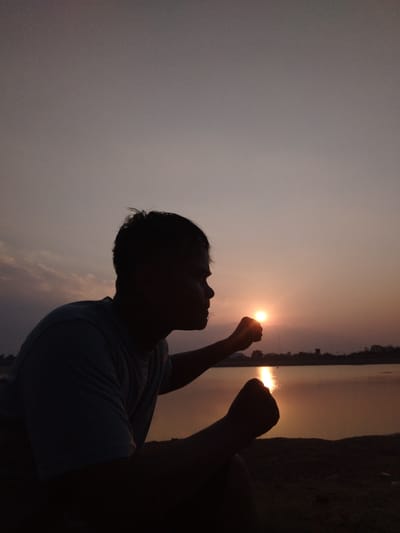 A silhouette of a person is positioned in the foreground, facing right, with their head tilted slightly. The person's right hand is extended forward, with their fist closed, as if holding or interacting with the setting sun. The left hand is also clenched in a fist, held closer to the body. The background features a body of water reflecting the warm hues of the sunset. A faint treeline is visible on the horizon, beyond the water. The sky is a gradient of soft purples and oranges, with some wispy clouds. The overall lighting suggests it is dusk or dawn, with the sun low on the horizon. The location is identified as Sambirejo, Indonesia.