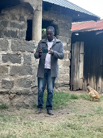 The image displays a man standing in an outdoor setting in Kiganjo, Kenya. The man, with a dark complexion, is dressed in a grey long coat over a light checkered shirt, blue jeans, and dark shoes. He is holding a dark green smartphone horizontally with both hands, looking down at the screen, appearing focused on its content, possibly taking a picture or video.

He stands in front of a rustic stone building constructed from dark, rough-hewn blocks. Parts of a corrugated metal roof are visible above and to the left of the man, attached to the stone structure. Wooden planks are also incorporated into the upper section of the stone wall. The ground around him is a mix of sparse grass and dirt.

To the right and slightly behind the man, a brown and white chicken is seen pecking at the ground, foraging for food. The lighting suggests it is daytime, with soft illumination, possibly from an overcast sky or early morning/late afternoon, as there are no harsh shadows. No specific emotions are discernible on the man's face beyond concentration, and no visible text appears in the frame.