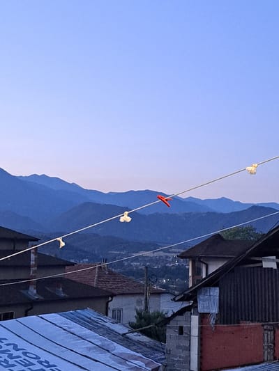This image captures a scene in Simitli, Bulgaria, featuring a twilight sky with mountains in the background. In the foreground, clotheslines stretch across the frame, adorned with several clothespins. One red clothespin is prominently visible, holding what appears to be a piece of fabric or paper. Other clothespins hold small, light-colored objects, possibly also fabric. The scene is set against a backdrop of residential buildings with tiled roofs, and hints of the town of Simitli are visible in the mid-ground. The overall impression is one of a quiet evening in a Bulgarian town, with laundry drying in the open air against a scenic mountainous landscape.