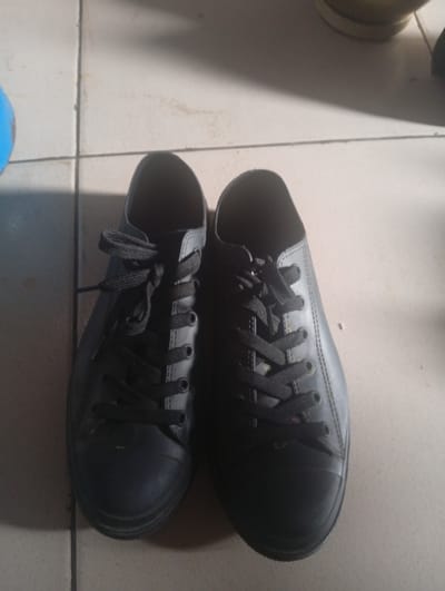 A pair of black, low-top lace-up sneakers is shown resting on a tiled floor. The shoes are positioned side-by-side, with the laces tied in bows. The floor is composed of light-colored square tiles with visible grout lines. The lighting suggests an indoor environment, possibly a room or hallway. The specific location in Ashata, Nigeria, is not discernible from the image itself.