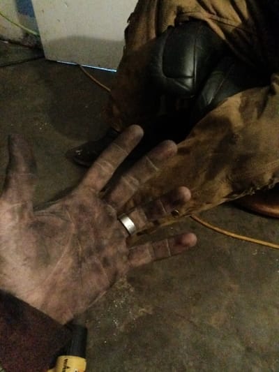 A dirty hand, wearing a silver ring, is displayed against a background of worn brown leather and black knee pads. The hand appears grimy with dark smudges across the palm and fingers. The leather garment nearby seems equally weathered. A dark boot with a buckle rests on the floor, alongside a yellow electrical cord. The overall scene suggests a work environment or a setting involving manual labor.