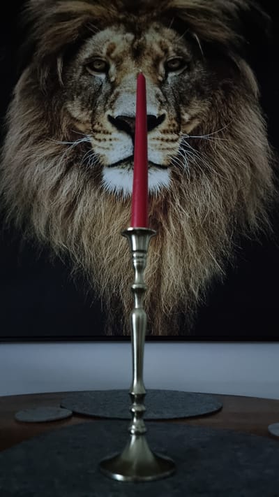 A red candle stands upright in a brass candlestick, positioned in the foreground of the image. The candle is centered and appears to be the primary subject. Behind the candlestick and candle is a framed artwork depicting the face of a lion. The lion's mane is prominent and detailed, taking up a significant portion of the background. The artwork is mounted on a wall, the top edge of which is visible in the upper portion of the image. The candlestick rests on a table surface, partially obscured by what appear to be two circular placemats or coasters. The lighting is dim, casting shadows and creating a somber atmosphere. The scene suggests an indoor setting, likely a home environment. There is no indication of the city of Heidenheim an der Brenz, Germany.