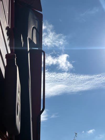 The image shows the underside of a red vehicle, possibly a truck or SUV, against a bright blue sky with wispy white clouds. The vehicle's interior is visible, including a black sun visor with a textured surface and a part of the vehicle's frame. A metal handle extends from the frame. A small branch with green leaves is partially visible in the lower left corner. The lighting suggests it is daytime, with the sun casting shadows on the vehicle's interior. The scene is outdoors, under a clear sky with some scattered clouds, indicating fair weather. There is no visible text in the image. The orientation of the image is such that the sky appears below the vehicle.