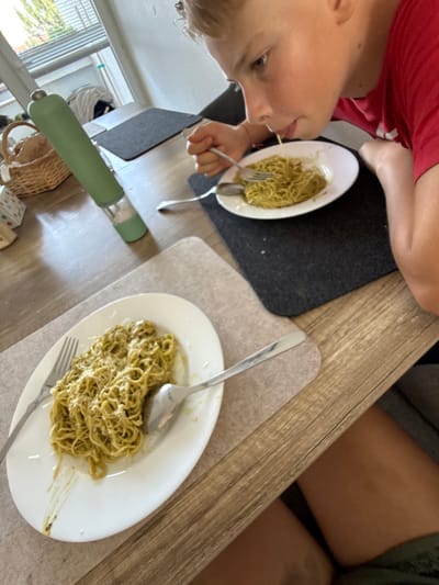 A young boy in a red shirt is eating pasta with a fork, his mouth close to the plate. The pasta is a light green color, likely pesto, and is served on a white plate. Another plate of the same pasta is visible on the table, along with a fork and spoon. A green thermal bottle and a woven basket are also on the table. The table is made of wood, and placemats are used. In the background, a window with blinds is visible, showing a glimpse of trees outside. The scene appears to be an indoor dining area, possibly a home. The image does not provide any specific clues to determine its location in Bruchköbel, Germany.