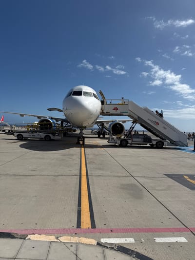The image displays a white passenger aircraft, consistent with an Airbus A320 series, parked on a concrete tarmac at an airfield in Granadilla de Abona, Spain. The aircraft is positioned head-on to the viewer. Its front port-side cabin door is open, with a mobile airstair, marked "AVIAVIATION SUPPORT" and "AP," connected to the opening. At least two individuals are visible on the airstair. The cowling of the aircraft's left engine is open, exposing internal machinery. Numerous pieces of ground support equipment, including what appear to be cargo loaders and maintenance platforms, are situated around the aircraft's wings and fuselage; some equipment is branded "AP" and "TDI airport." A tow bar is attached to the nose landing gear. Several indistinct figures, likely ground personnel, are present on the tarmac around the aircraft. The concrete tarmac features yellow and black centerline markings extending into the foreground, with red and white boundary lines present closer to the camera. A blue painted area is visible on the tarmac beneath the far end of the airstair. The scene is illuminated by direct sunlight, casting shadows towards the left, under a clear blue sky with scattered small white clouds.