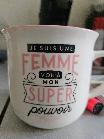 The image displays a white ceramic mug, slightly discolored with light brown residue along its pink rim, resting on a textured light-colored surface. The mug features a French inscription in various fonts and colors. The top line, "JE SUIS UNE," is rendered in white sans-serif capital letters on a black rectangular background. Below this, "FEMME" is prominently displayed in large, pink, sans-serif capital letters. Following, "VOILÀ" appears in white sans-serif capital letters on a black rectangular background, flanked by decorative black swirls. Underneath, "MON" is in black sans-serif capital letters. The next line, "SUPER," is in large, pink, sans-serif capital letters, enclosed within a black outlined banner design. Finally, "pouvoir" is written in black cursive script below the banner. To the right of the mug, partially out of focus, a small, cylindrical red object with a black end is visible on the surface. The background is uniformly blurred, suggesting an indoor setting. This scene is located in Villeneuve-Saint-Georges, France.