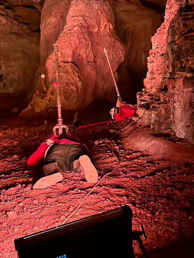 In Gollagutta, India, a person lies prone on a rocky, reddish ground within a dimly lit cavern. The individual, wearing a red jacket and dark pants, has their head turned away from the camera. Two tall, ornate staffs, possibly decorative or functional, stand nearby. One is positioned directly above the person's head, and another is further back. A red backpack is visible near the second staff. The cavern walls are rough and textured, illuminated by red lighting, suggesting a theatrical or controlled environment rather than a natural cave. The ground is uneven, covered in small stones and larger rocks. A black object, possibly a piece of equipment like a light or camera, is in the foreground, partially obscuring the lower portion of the image.