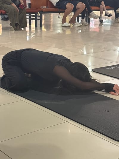 In Tri Protoka, Russia, a person is performing a yoga pose, specifically Balasana (child's pose), on a black yoga mat. The individual is wearing black athletic pants and a black top with sheer sleeves, and their arms are extended forward. The yoga mat is positioned on a tiled floor with light-colored square tiles. In the background, a row of orange seats is visible, with several individuals seated on them. One person on the left is wearing olive-green pants and brown shoes. Another person is wearing shorts. Other individuals in the background are wearing various footwear, including white sneakers and black shoes. The lighting suggests an indoor setting, possibly a public waiting area or a communal space.