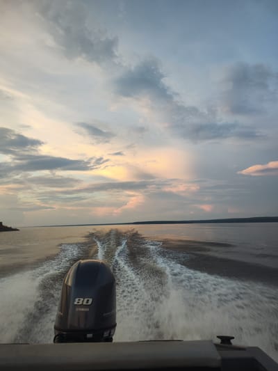 An outboard motor, labeled "80" and "YAMAHA", is visible from the stern of a boat, creating a wake on a calm body of water. The wake extends behind the boat, forming a pattern of white spray and ripples. The scene appears to be set at dusk, with the sky displaying a gradient of colors from pale yellow and pink near the horizon to a muted blue overhead. Clouds are present in the sky, with some appearing darker and more dramatic than others. The horizon is visible in the distance, with a faint line of land or trees. The location is identified as Galewo, Russia.