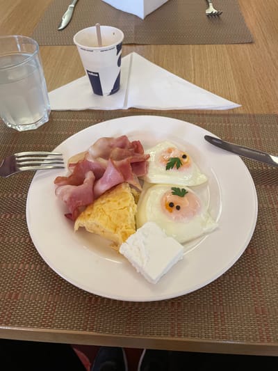 The image shows a breakfast plate on a table setting. The plate contains two fried eggs, decorated with parsley and black pepper to resemble faces, along with bacon, scrambled eggs, and a piece of white cheese. Cutlery, including a fork and knife, are placed around the plate. A glass of water, a coffee cup with a stirrer, and a napkin are also present on the table. The table has a woven placemat and a wooden surface. The photo was likely taken indoors in Otopeni, Romania.
