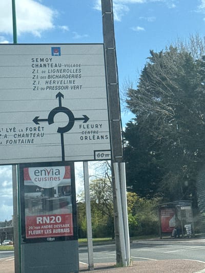 An outdoor scene in Fleury-les-Aubrais, France, captures a multi-directional road intersection under a partly cloudy blue sky. In the foreground, a large white directional sign is mounted on a grey support pole. The upper section of the sign, bearing a small red and blue pictogram, indicates straight-ahead routes to "SEMOY", "CHANTEAU-VILLAGE", and various industrial zones (Z.I.). Below this, a graphic depicts a roundabout, with a left arrow pointing towards destinations including "ST LYÉ LA FORÊT", and a right arrow indicating "FLEURY", "CENTRE", and "ORLÉANS". A "60 m" distance marker is visible to the right of the roundabout graphic.
Directly beneath the road sign, a vertical illuminated advertising panel displays an "envia cuisines" advertisement featuring kitchen interiors and appliances. The bottom section of this panel specifies "RN20" and the address "20, rue ANDRÉ DESSAUX FLEURY LES AUBRAIS", with a "Cityz" logo at the very bottom.
To the right of the signs, a dense cluster of trees, primarily dark green conifers and leafless deciduous trees, occupies the midground. Further to the right, a transparent-roofed bus stop shelter is situated beside the road. Inside the shelter, a single individual, dressed in dark clothing, is seated, partially obscured by an internal red advertisement and a stack of what appears to be newspapers or magazines beside them. The scene is bisected by an asphalt road with visible lane markings and a textured roadside surface. A light-colored car is partially visible in the distant background on the left.