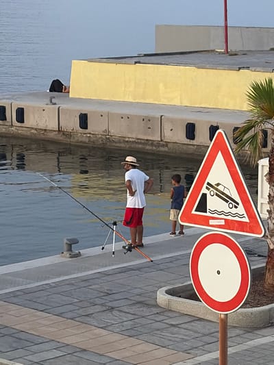 This media file depicts a waterfront scene in Imperia, Italy. The primary subjects are two people: a man fishing and a child. The man, wearing a hat, white shirt, and red shorts, is actively fishing with a rod and reel set up on a tripod. The child, in a blue shirt and light shorts, stands behind him, observing.
The setting is a paved walkway next to the water, likely a harbor or marina. A yellow building is visible in the background, along with some concrete structures. There are two prominent traffic signs. The first is a triangular warning sign with a red border and a white interior, displaying a black pictogram of a car tilting into the water, indicating a drop-off or submerged edge. The second is a circular prohibition sign, also with a red border and a white interior, featuring a black dot in the center, which typically signifies "no stopping" or "no entry" depending on context and accompanying signs.
The lighting suggests it is daytime, possibly late afternoon, with the sun casting shadows. The water appears calm, reflecting the surrounding structures and sky. The overall atmosphere seems peaceful, with the focus on the activity of fishing. There is no visible text on the clothing or other elements in the image.