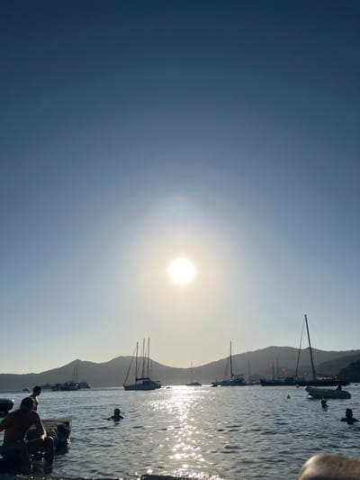 In the waters of the city of Sant Josep de sa Talaia, Spain, a bright sun shines in a clear blue sky, casting a shimmering reflection on the sea. The sun is positioned slightly to the left of the center of the frame, creating lens flares. Several sailboats of varying sizes are anchored in the bay, silhouetted against distant, hilly landmasses. In the foreground, people are in the water. On the left, a man sits on a stone structure partially submerged in the water, with another man standing behind him. Two other individuals are visible in the water further out, their heads above the surface. The overall scene depicts a tranquil day at the beach with calm waters and several boats.