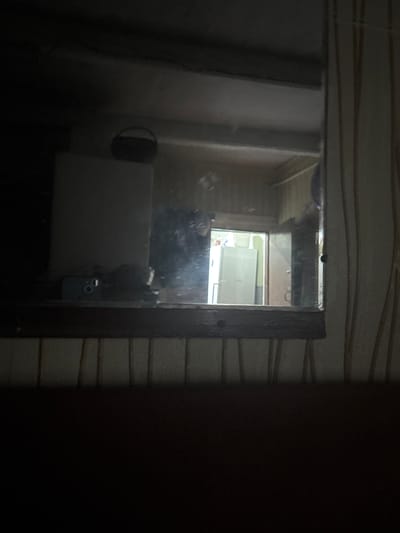 The media file is a photograph taken in a dimly lit room, likely a bathroom. The primary subject is a mirror reflecting a portion of the room. In the reflection, a light-colored appliance, possibly a washing machine or dryer, is visible. Above it, a window with a white frame allows some light to enter, illuminating a part of the room. The mirror itself appears to have some smudges or watermarks. The surrounding wall has a textured pattern, possibly tiles with horizontal lines. The lighting is low, suggesting it might be nighttime or the room is not well-lit. A dark, indistinct object is also visible in the foreground, partially obscuring the mirror, and a dark vertical frame of the mirror is present on the right side of the image. The location context provided is Ганчарскі сельскі Савет, Belarus, which is a rural council area. There is no visible text in the image. The scene evokes a sense of a domestic, perhaps slightly dated, environment.