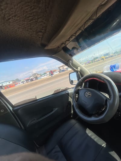 The image is taken from inside a vehicle, looking out towards a busy street scene. The perspective is skewed, making it appear as if the vehicle is tilted.
**Subjects:** The primary subjects are the interior of the vehicle, including the steering wheel, dashboard, and part of a black leather seat. Outside, a street lined with numerous people and various types of vehicles is visible. These vehicles appear to be minibuses or similar small passenger transport, some brightly colored like orange and white, others more utilitarian. There are also market stalls and vendors set up along the roadside, indicated by colorful umbrellas and makeshift structures.
**Activity or Event:** The scene depicts a bustling marketplace or a busy transport hub. People are gathered, likely engaged in commerce or waiting for transportation. The numerous vehicles suggest a flow of people moving in and out of the area.
**Notable Details:**
* **Time of Day:** The bright sunlight and clear blue sky indicate it is daytime, likely mid-morning or afternoon.
* **Weather:** The weather appears sunny and clear.
* **Location Cues:** The presence of many minibuses, market stalls, and the overall vibrancy of the scene are consistent with a busy urban or semi-urban area in Nigeria, specifically Mowe. A water bottle with a blue label is visible on the dashboard, with text that includes "CLAY." There's also a partial phone number visible on the side of a building or vehicle: "070757964."
* **Emotions:** The scene evokes a sense of lively activity and commerce.
**Visible Text:** "CLAY" is visible on a water bottle. A partial phone number "070757964" is visible. The word "AIRBAG" is visible on the steering wheel cover.