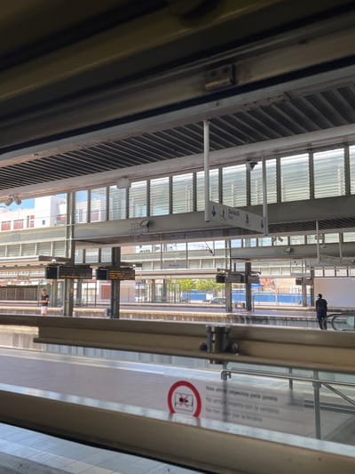 This image captures a view from inside a train at a station in Aveiro, Portugal. The perspective is looking out through a window, showing the platform and the tracks. Visible are signs with text in Portuguese and possibly other languages, one of which reads "Saída Exit" with an arrow pointing left, indicating an exit. Another sign, partially obscured, contains instructions in multiple languages, including "Não atirar objectos pela janela" which translates to "Do not throw objects out the window." A circular red and white icon with a diagonal line across an object being thrown suggests a prohibition.
In the background, other trains are visible on adjacent tracks, and parts of the station's architecture, including a glass-covered roof and metal support structures, are apparent. A few people are visible on the platform and inside another train, one person is standing and looking out, another appears to be walking away. The scene suggests a typical day at a train station, likely during daylight hours, with no specific weather conditions being particularly noticeable. The overall atmosphere is one of transit and everyday activity.