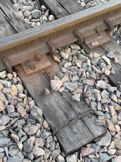 The image shows a close-up, overhead view of a section of railroad tracks. The primary subjects are the metal rails, wooden railway ties, and the gravel ballast that supports the tracks. The scene is outdoors, with the gravel composed of various shades of grey, pink, and tan rocks. The wooden ties appear weathered and worn, with visible grain and some splintering. The metal components, including the rail and fastening clips, are rusted and textured.
There is no visible activity or event taking place; the image captures a static, detailed view of the railway infrastructure. Notable details include the way the gravel fills the spaces between the ties and around the rails, suggesting a functional railway line. The lighting appears to be natural daylight, but there are no specific cues to determine the time of day or weather conditions. The location context of Kant, Kyrgyzstan, is provided but not visually evident in the image itself. There is no visible text within the image. The overall impression is one of rugged, industrial construction and the passage of time, conveyed through the textures and wear of the materials.
