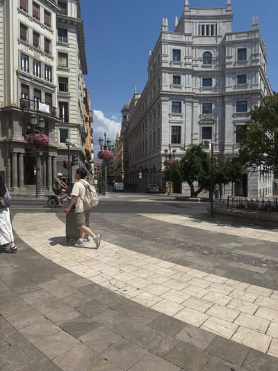 The image shows a street scene in Granada, Spain, likely in a historic district given the architecture. In the foreground, two individuals are walking on a paved sidewalk with a patterned design. The person on the left is wearing a long white skirt and sandals, and the person on the right is wearing a t-shirt, shorts, and a backpack, pulling a wheeled suitcase. They are walking in the same direction. Behind them, a man on a bicycle with a helmet is also cycling in the street.
The street is lined with large, ornate buildings that appear to be several stories tall. One building on the right is made of light-colored stone with classical architectural details, including columns and decorative elements around the windows. Another building on the left is also substantial, with multiple windows and balconies, and appears to be made of a lighter stone or stucco. Streetlights with hanging flower baskets are visible along the street, adding a touch of color.
The sky is a clear blue with some scattered white clouds. The sun is shining, casting shadows on the ground. The street itself is paved with large, rectangular stones in varying shades of gray and beige, with some darker, reddish-brown accents in a band. A crosswalk is visible in the middle ground, leading to another street. Traffic signals are present, indicating this is a functioning intersection. A dark-colored car is parked on the side of the street in the middle ground, and a white van is visible further down the road. Trees with green foliage are planted along the sidewalk, contributing to the urban landscape. The overall impression is a sunny day in a European city with a blend of historic and modern elements.